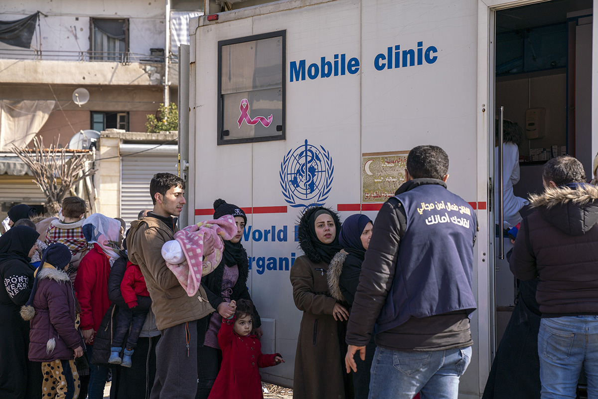 Caregivers with their children at a UNICEF-supported mobile health clinic waiting for consultations, nearby a makeshift shelter, in Aleppo city, north Syria. © UNICEF/ Muhannad Al-Asadi Adults and children stand in line a mobile clinic