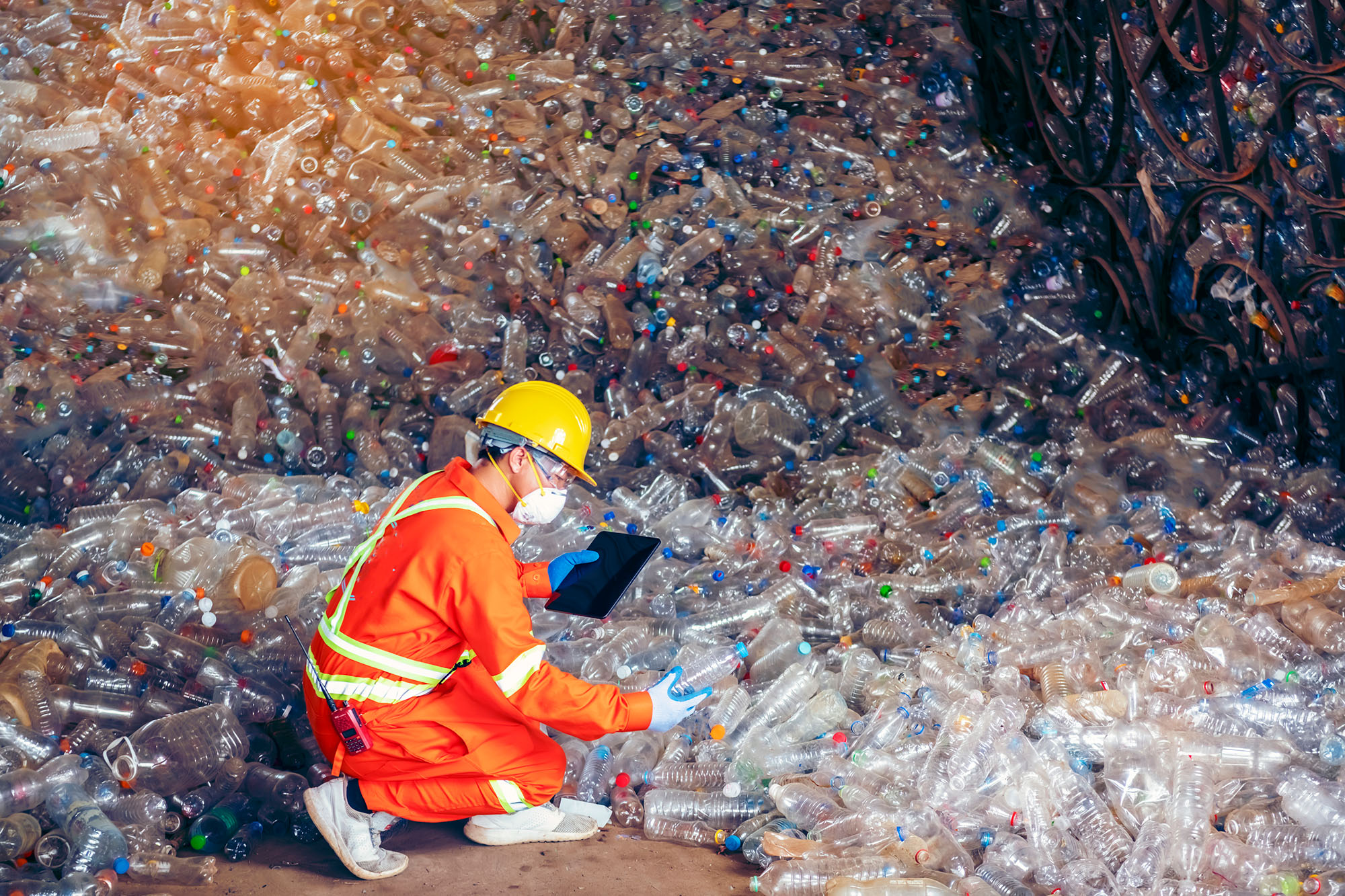 A man holding a tablet in front of a huge pile of empty plastic water bottles.
